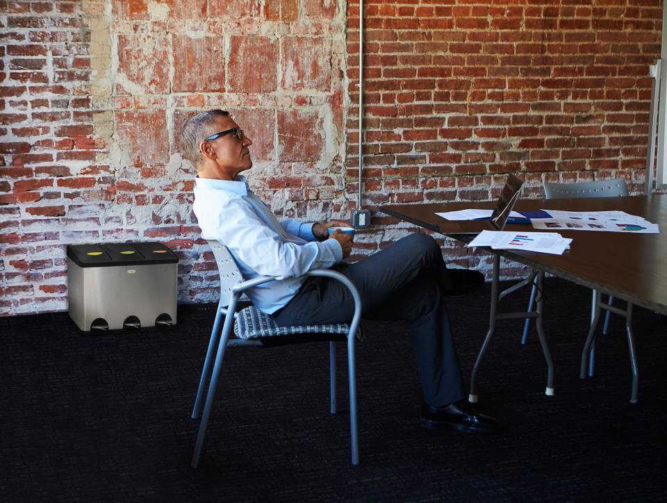Un homme au bureau se détend sur une chaise devant une table avec un ordinateur portable et des documents, un mur de briques en arrière-plan.