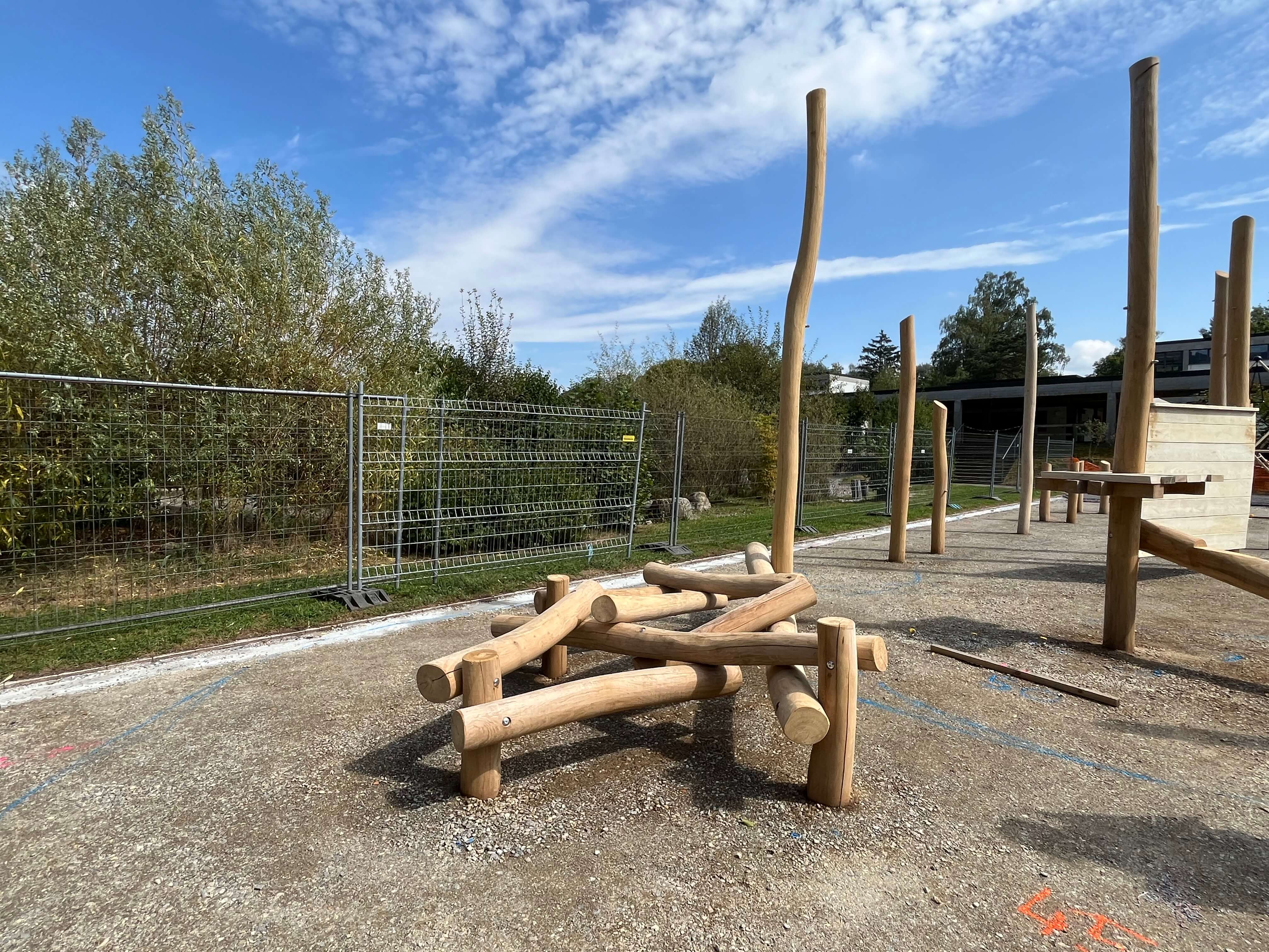 Bild eines im Bau befindlichen natürlichen Spielplatzes mit Holzstrukturen. Im Hintergrund sind Bäume, ein Zaun und ein blauer Himmel zu sehen.