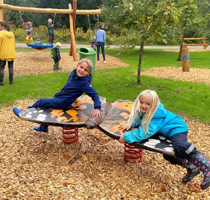 Zwei Kinder haben Spaß auf einem Wipptier in einem Park. Im Hintergrund sind andere Kinder und Erwachsene beim Spielen und Spazieren zu sehen.