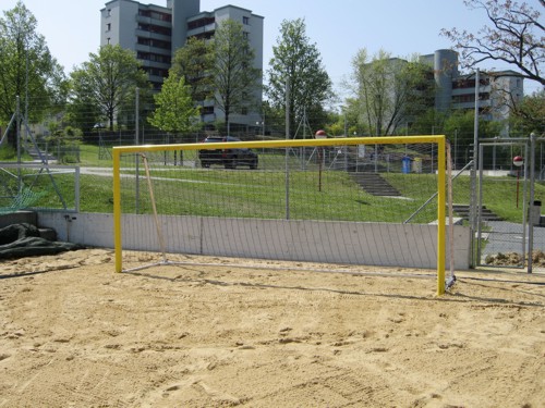 Gelbes Fußballtor auf einem sandigen Spielfeld, umgeben von grünen Bäumen und Wohnhäusern im Hintergrund