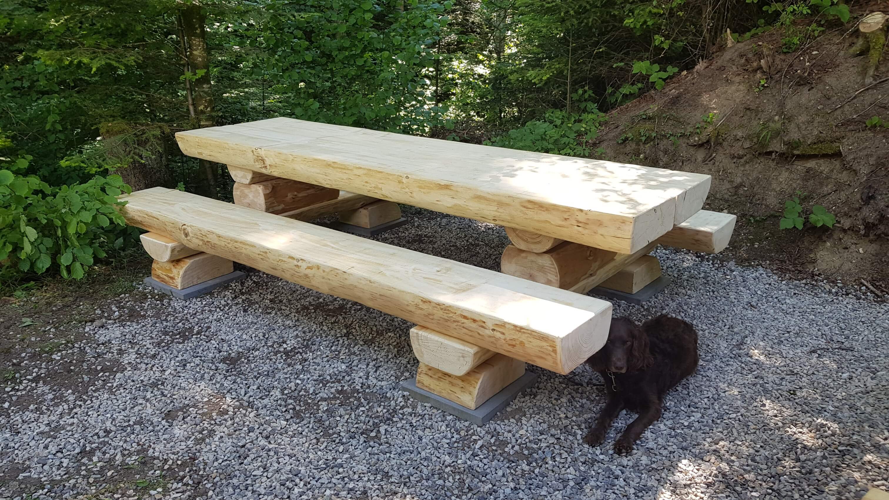 Holztisch und -bänke im Freien auf Kiesboden, umgeben von Bäumen