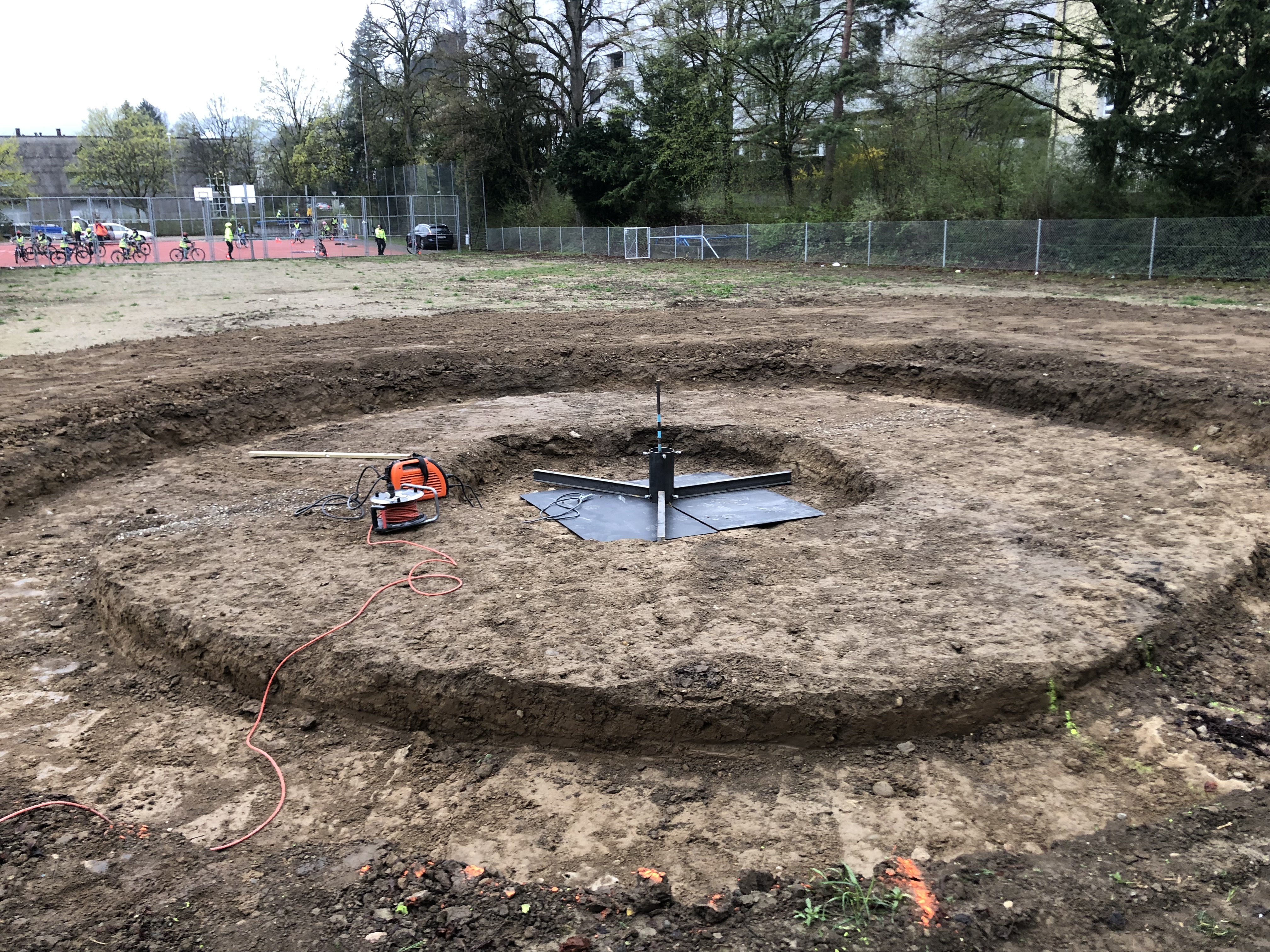 Ein ausgehobenes Erdloch auf einer Baustelle, das von einem Zaun umgeben ist, mit einem orangenen Werkzeug und Bauutensilien in der Mitte.