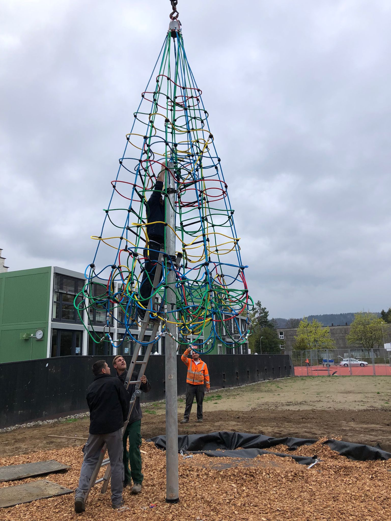 Mehrere Männer installieren ein buntes Klettergerüst auf einem Spielplatz. Einer steht auf einer Leiter und arbeitet am Gerüst, während andere den Prozess überwachen. Im Hintergrund sind Schulgebäude und Bäume sichtbar.