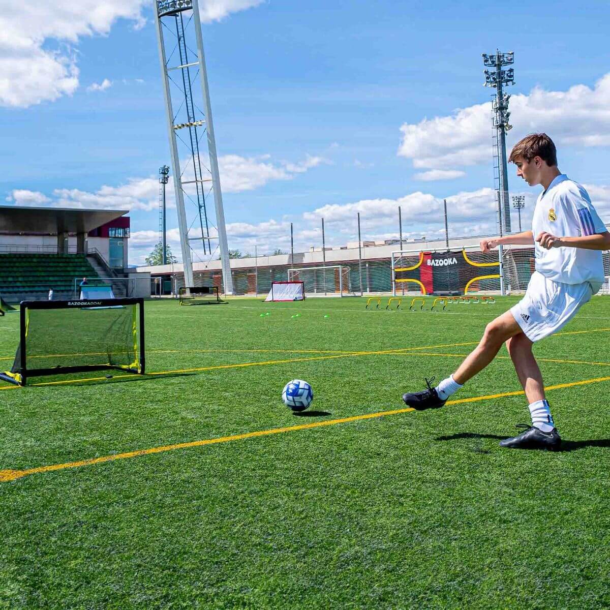 Ein junger Mann in weißen Sportkleidung spielt Fußball auf einem grünen Rasenplatz mit kleinen Toren und Übungshürden im Hintergrund