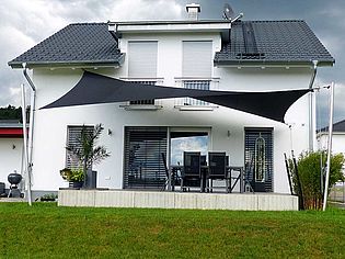 Vue de face d'une maison individuelle moderne de deux étages avec voile d'ombrage noir sur la terrasse et jardin soigné