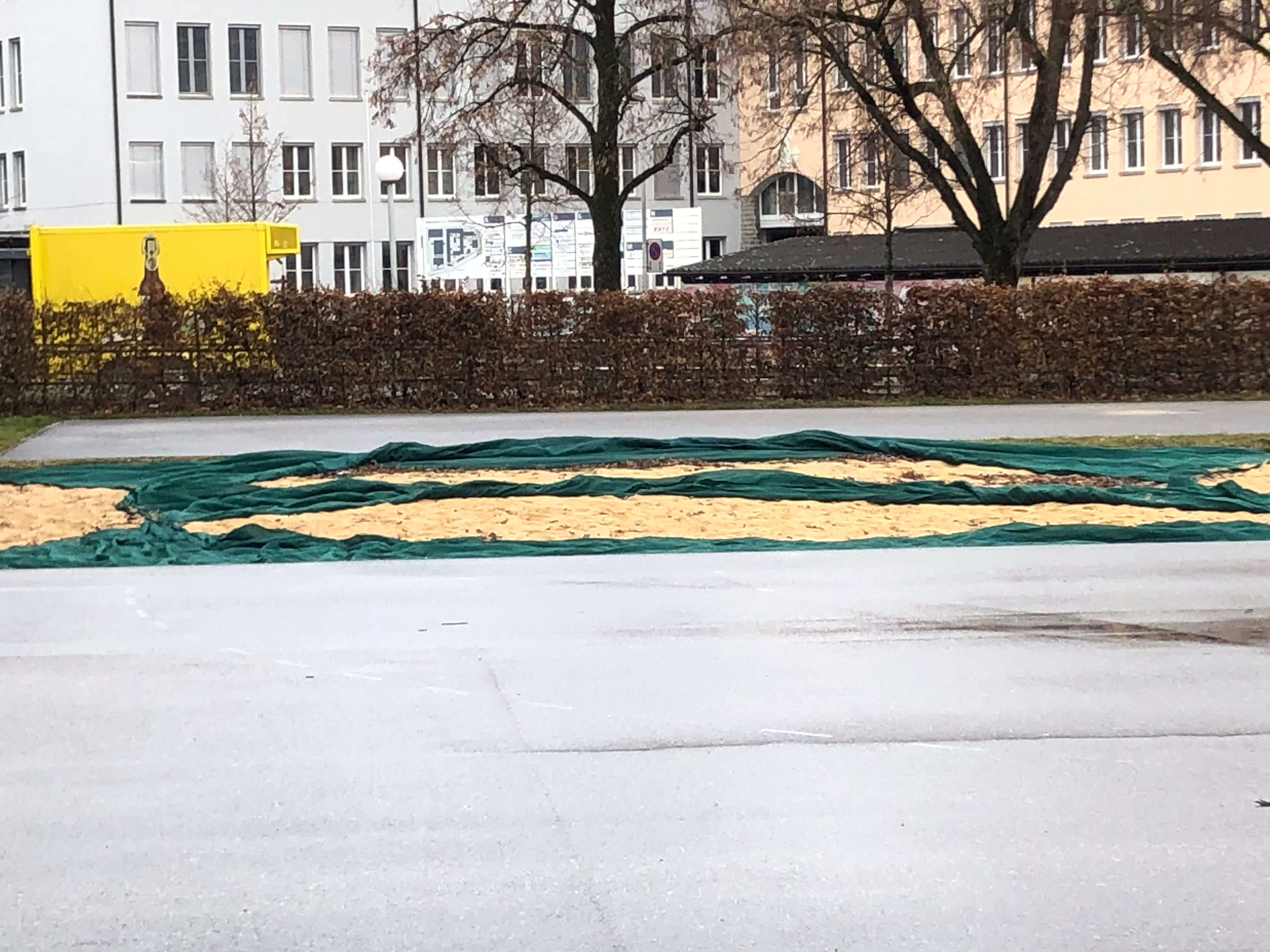 Sandfläche mit grüner Plane bedeckt, umgeben von Gebäuden und Bäumen