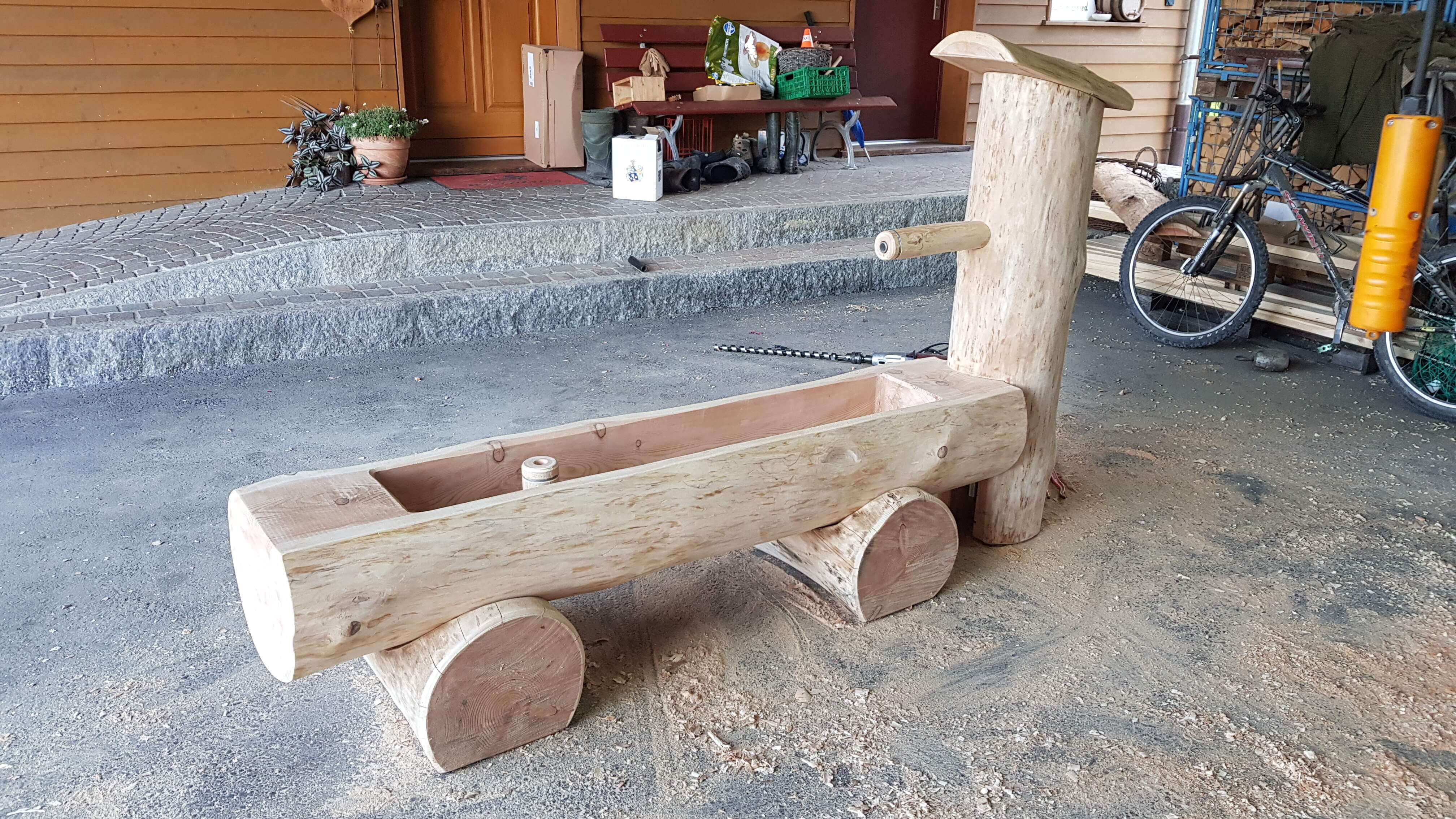 Une fontaine en bois rustique, fabriquée à la main à partir de troncs d'arbres, se trouve dans une cour pavée. En arrière-plan, on peut voir différents outils de jardinage.