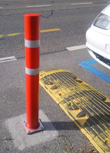 Roter Absperrpfosten auf einem Bürgersteig neben einem gelben Bordstein und einer Straße.