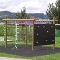 Une structure d'escalade pour aire de jeux avec un mur de blocs et une corde à grimper, entourée d'un paysage vert et d'arbres en arrière-plan.