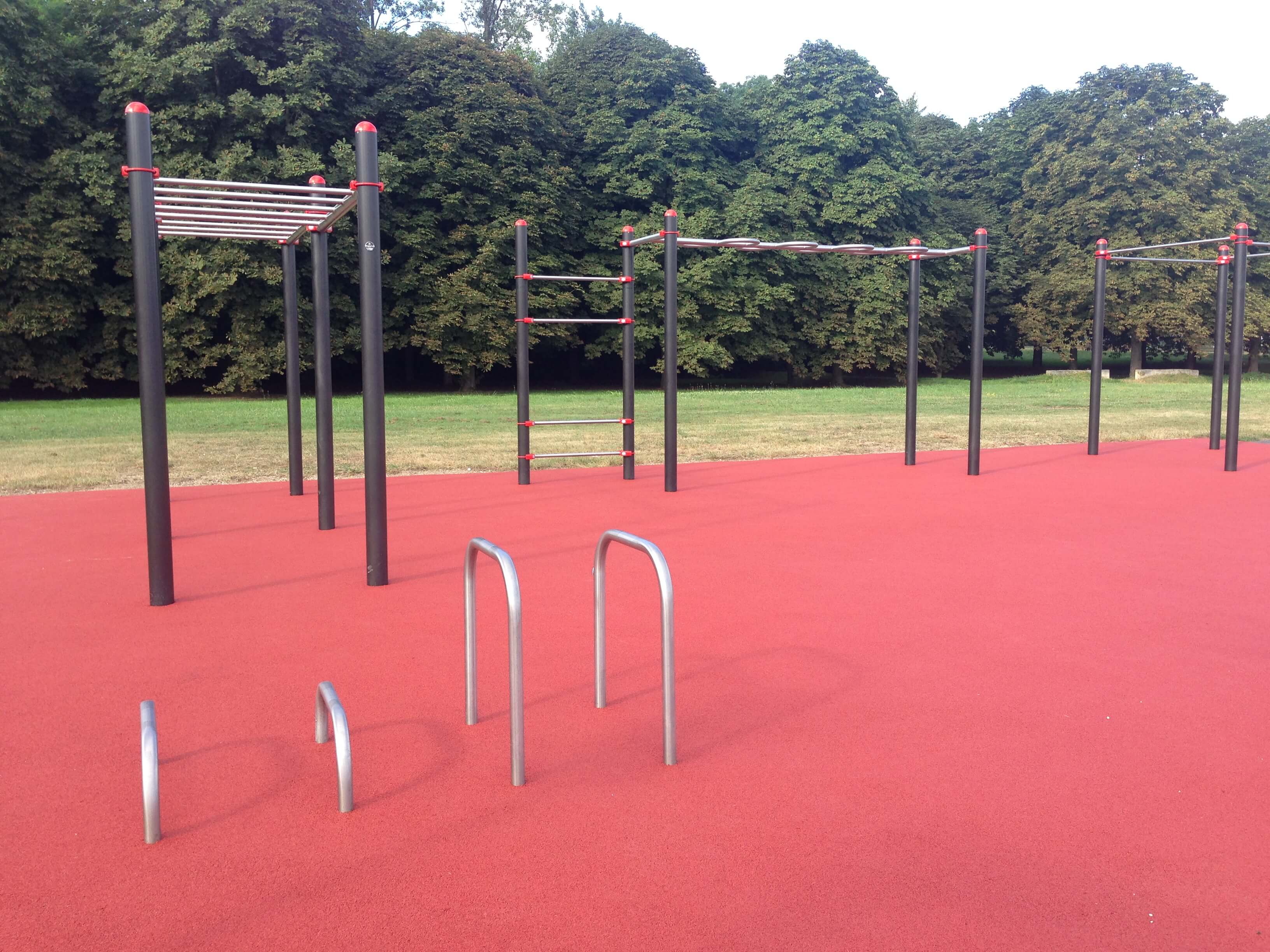 Un parc de fitness extérieur moderne avec différents appareils d'escalade sur un revêtement de sol rouge, entouré d'arbres verts en arrière-plan.