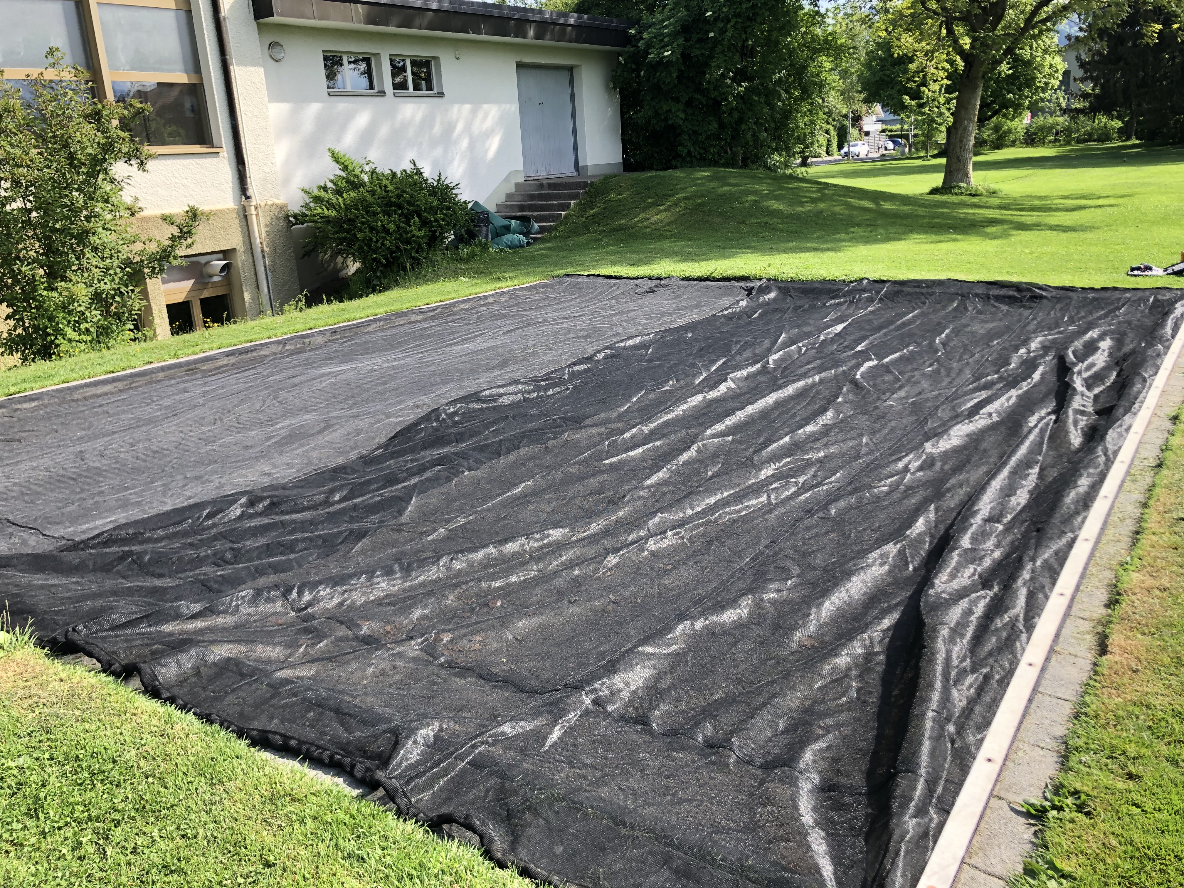 Großes Unkrautvlies, das als Abdeckung für ein Beet in einem Garten verwendet wird, neben einem Gebäude und umgeben von Rasen und Bäumen.
