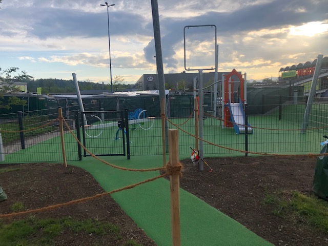 Une aire de jeux moderne en plein air avec portique, toboggan et revêtement de sol vert, entourée d'une clôture.
