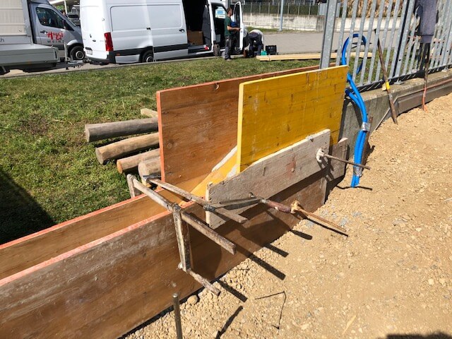 Murs de coffrage en bois pour des travaux de bétonnage sur un chantier, avec des véhicules de livraison garés en arrière-plan et une pelouse.