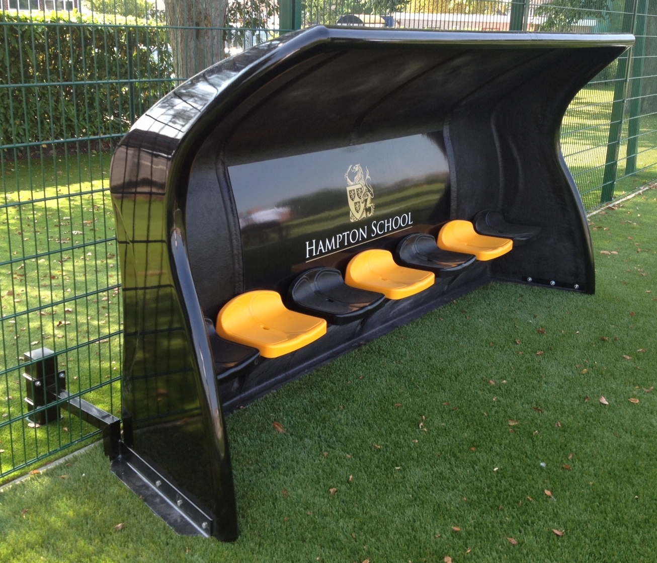 Schwarze Mannschaftsbank mit gelben Sitzen auf grünem Sportplatz der Hampton School, umgeben von einem Metallzaun.