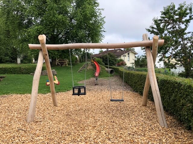 Une aire de jeux accueillante avec balançoire en bois, toboggan et base en copeaux de bois, entourée d'arbres et de pelouse.