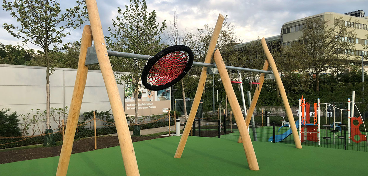 Ein moderner Spielplatz mit einer Schaukel, die aus Holzpfosten und einem Netzsitz besteht, umgeben von Bäumen und einer grünen Spielfläche.