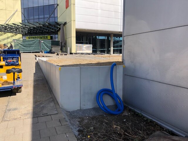 Un chantier de construction avec une rampe, des conteneurs de chantier et des tuyaux bleus devant un centre commercial.