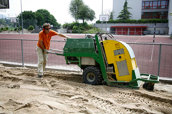 Un homme portant un T-shirt orange utilise une machine pour nettoyer le sable d'un terrain de sport en plein air