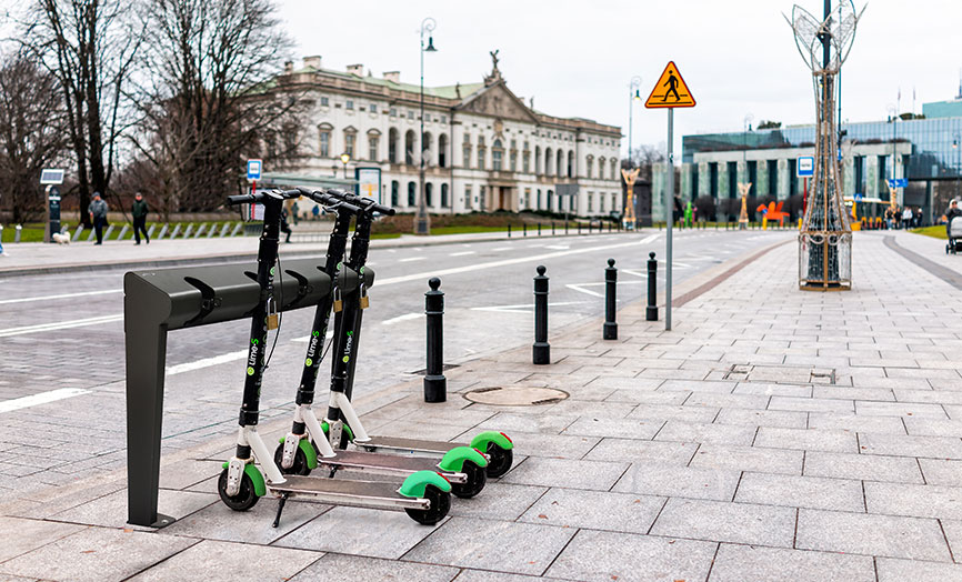 Drei Elektroroller sind an einer Mietstation in einer Stadt geparkt, mit einer Straße und historischen Gebäuden im Hintergrund.