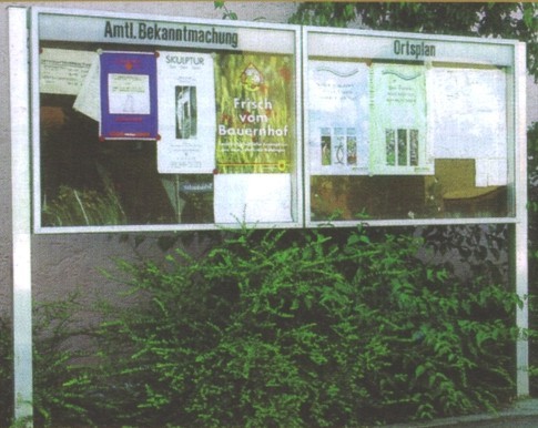 Eine öffentliche Anschlagtafel zeigt amtliche Bekanntmachungen und einen Ortsplan.