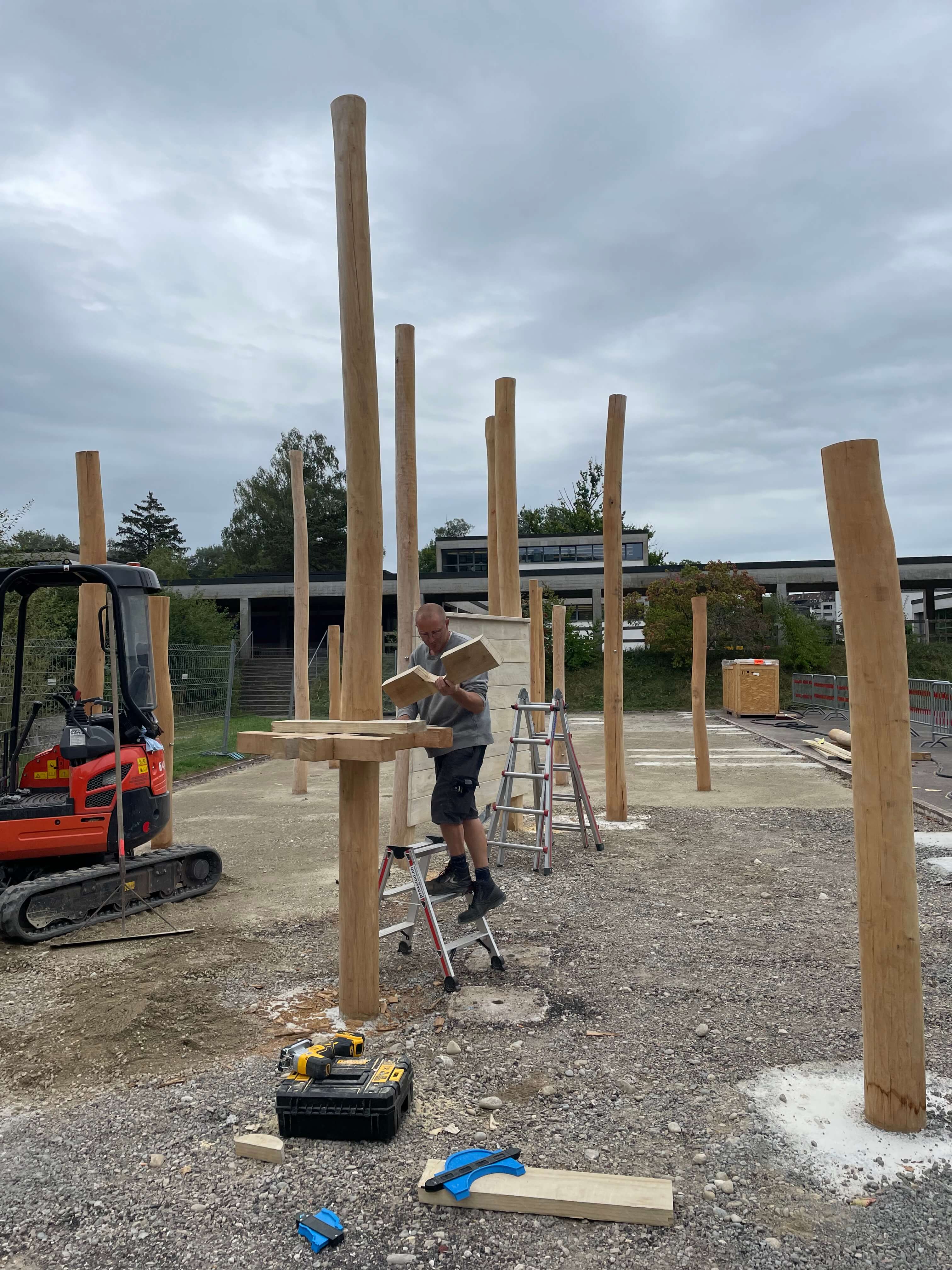 Ein Arbeiter steht auf einer Leiter und befestigt massive Holzpfosten auf einer Baustelle im Freien. Im Hintergrund ist ein kleiner Bagger zu sehen.