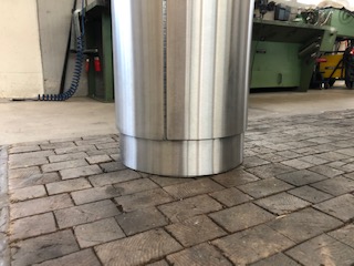 Un grand conteneur cylindrique en métal léger est posé sur un sol pavé dans un atelier. Des machines sont visibles à l'arrière-plan.