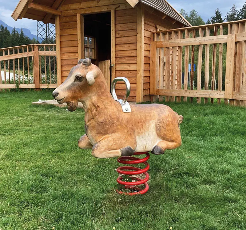 Eine Federwippe in Form einer Ziege auf einem grünen Rasen vor einem Holzhaus auf einem Spielplatz.