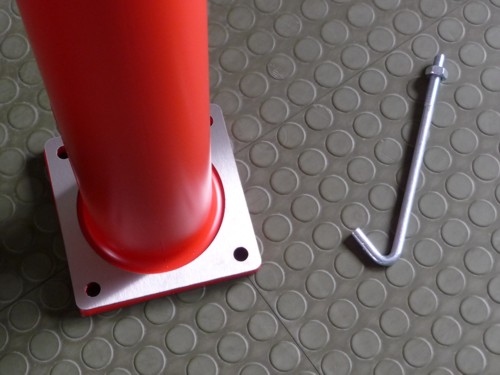 Nahaufnahme eines roten Metallpfostens mit quadratischer Bodenplatte, daneben liegt ein silberner Befestigungshaken auf einem strukturierten Bodenbelag.