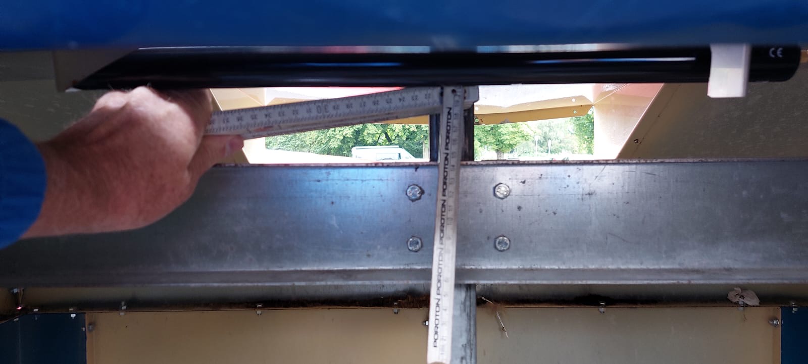 Ein Messschieber wird von einer Hand an einer Metallstruktur angebracht, um eine horizontale und vertikale Messung durchzuführen. Im Hintergrund sind Bäume und ein Fahrzeug sichtbar.