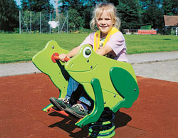 Une fillette souriante est assise sur un jouet à bascule à ressort en forme de grenouille sur une aire de jeux.