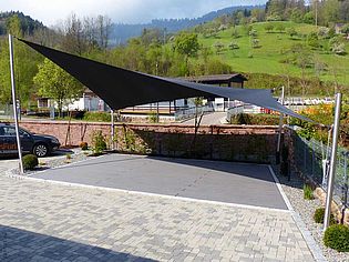 Une grande voile d'ombrage triangulaire offre de l'ombre dans une cour pavée avec un paysage verdoyant en arrière-plan.