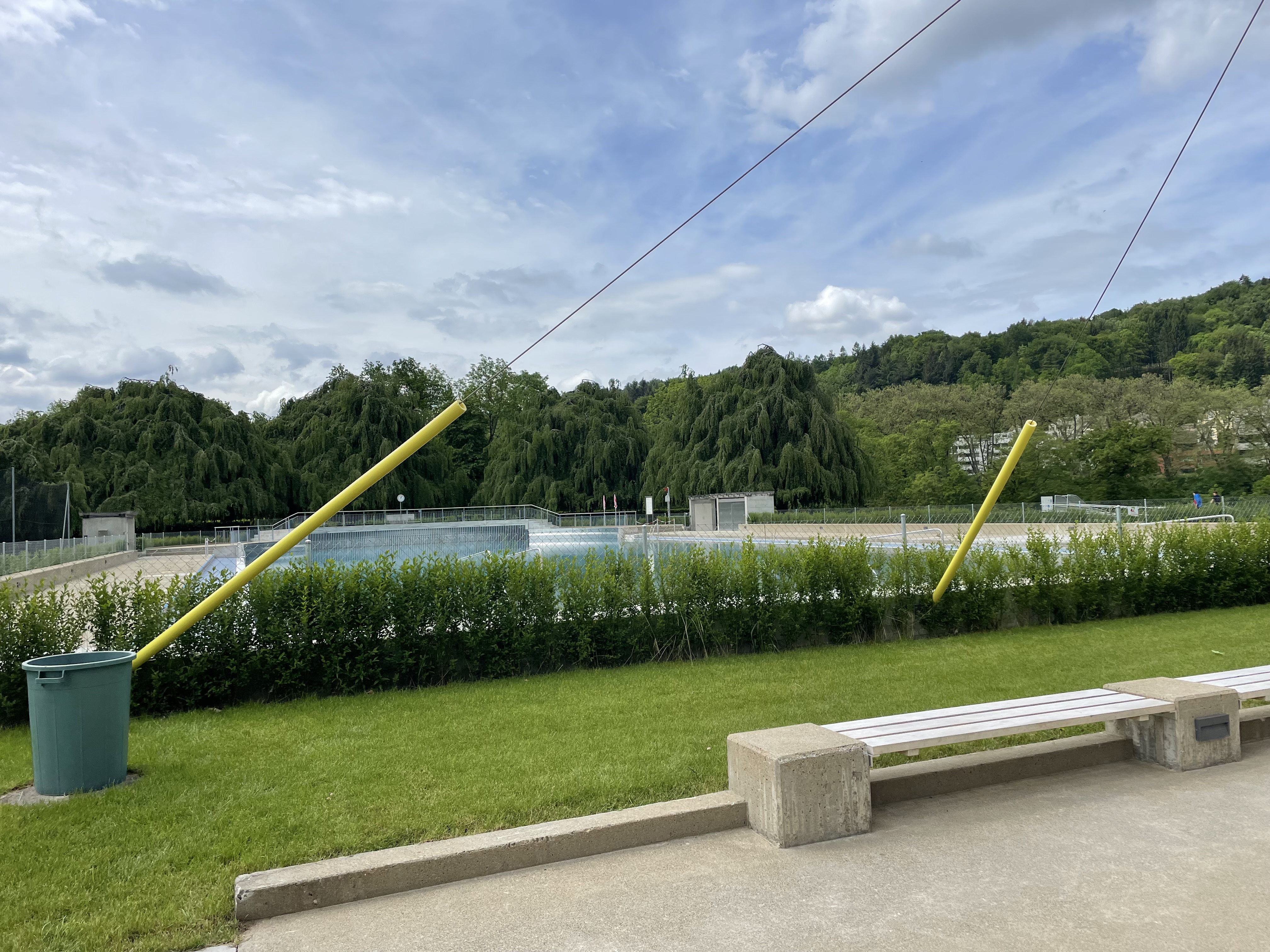 Ein leeres Freibad umgeben von grünen Bäumen und Wiesen mit einer Bank im Vordergrund unter bewölktem Himmel