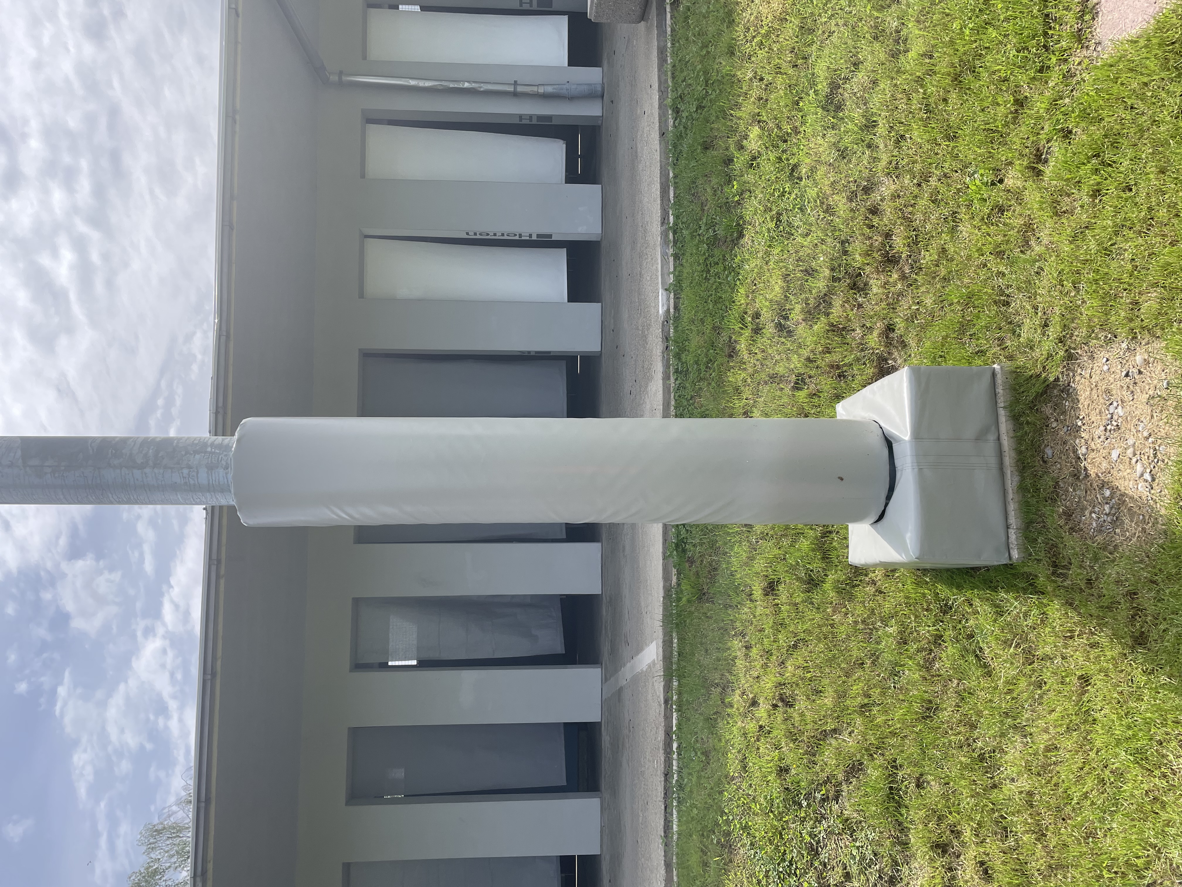 Ein Metallpfosten mit weißer Schutzhülle steht auf einer grünen Wiese vor einem Gebäude mit mehreren Türen.
