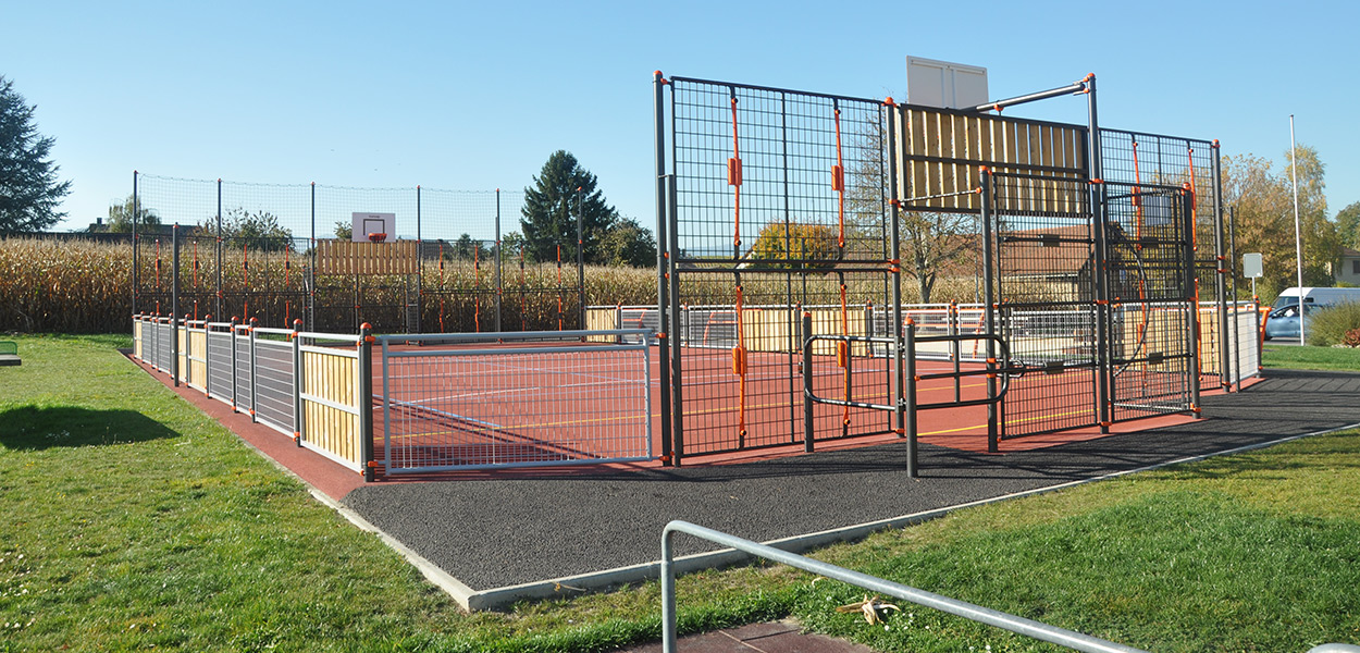 Ein eingezäunter, moderner Multifunktions-Sportplatz mit rotem Belag, geeignet für verschiedene Sportarten, unter blauem Himmel.