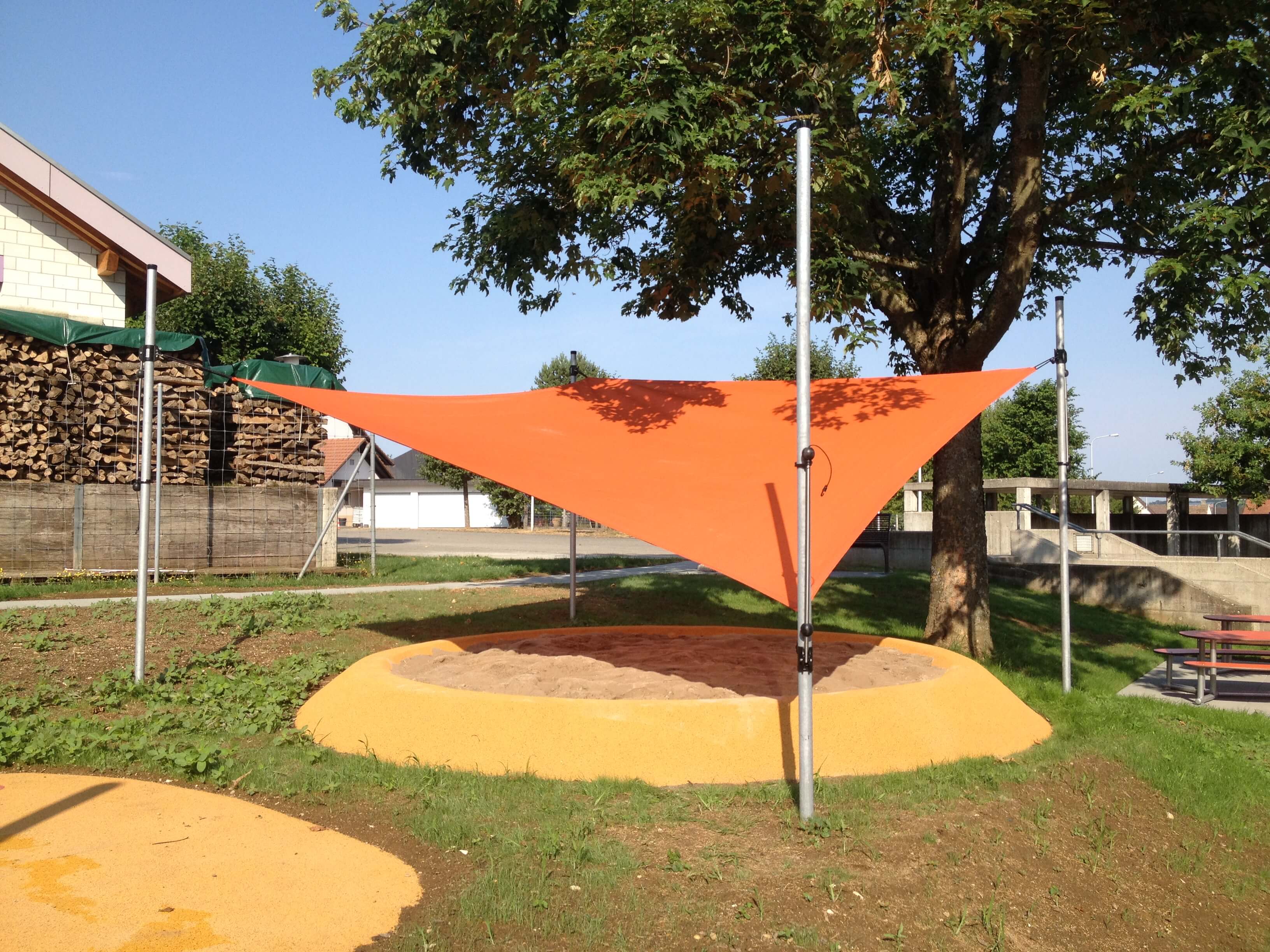 Un auvent orange offre de l'ombre au-dessus d'un bac à sable dans une aire de jeux, entouré d'arbres et de bâtiments.