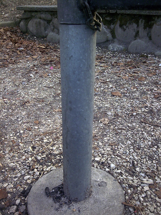 Ein einzelner Metallpfosten montiert auf einem Betonfundament, umgeben von Kies und Herbstlaub.