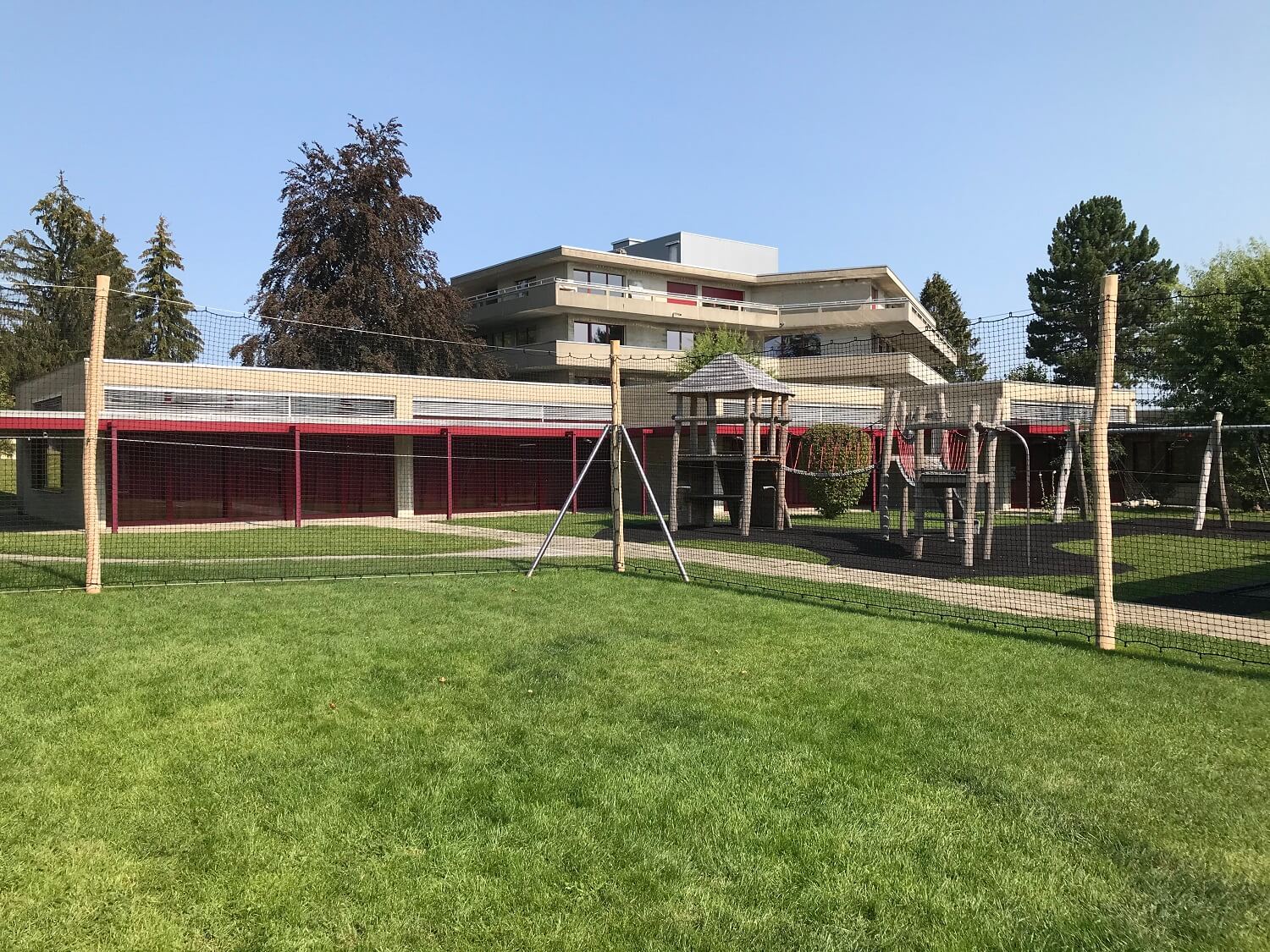 Une aire de jeux moderne avec portique et filet de sécurité sur une pelouse bien entretenue, entourée de bâtiments et d'arbres.