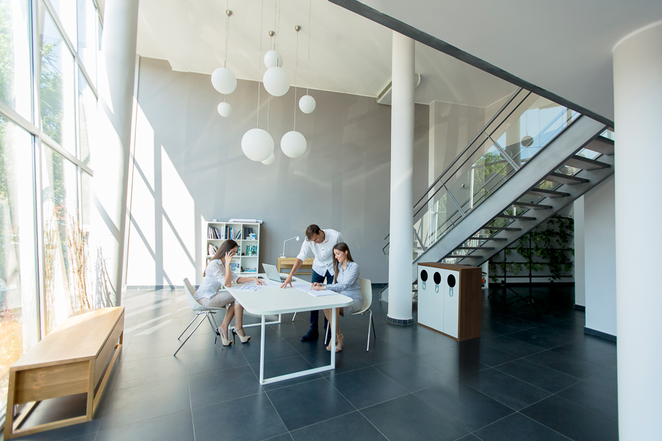 Helles, modernes Büro mit drei Personen, die an einem Tisch arbeiten, umgeben von großen Fenstern und einer modernen Treppe.